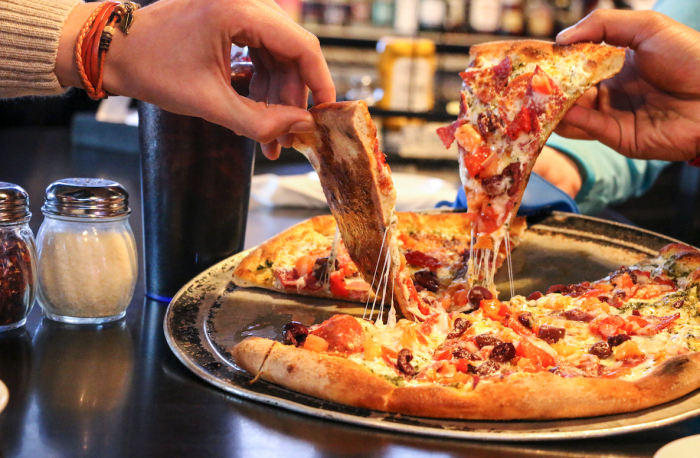 two people's hands each lifting out a slice of pizza from the whole pizza