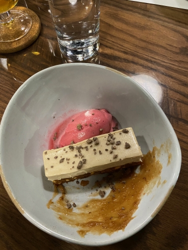 Brown rectangle of banana cake topped with white cream next to a pink scoop of sorbet and a smear of brown caramel - in a white bowl