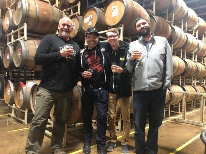 four men standing in front of racks of wooden barrels aging beer inside